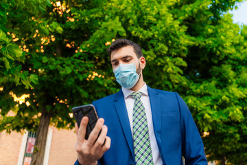 Young elegant businessman with surgical mask looks at phone in park