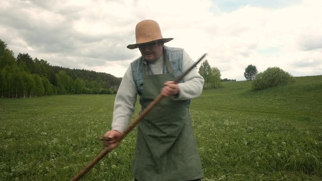 strong man 60-65 years old with a scythe mows green grass on the field. A male farmer in a hat works on the ground. Agriculture in the summer. A pensioner works in the village.