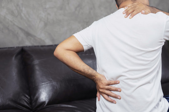 Man suffering touching his from back pain and shoulder sitting on sofa, grabbing a waist. Muscle spasm. Health care concept.