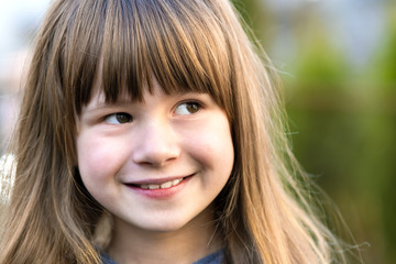 Portrait of pretty child girl with gray eyes and long fair hair smiling outdoors on blurred green bright background. Cute female kid on warm summer day outside.