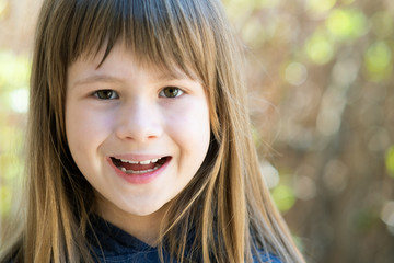 Fototapeta premium Portrait of pretty child girl with gray eyes and long fair hair smiling happily outdoors on blurred green bright background. Cute female kid on warm summer day outside.