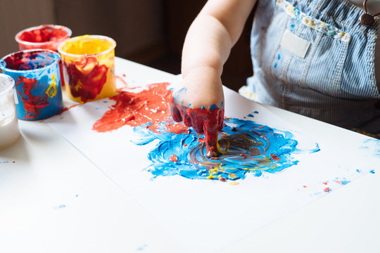 Child painting with her hands on the table at home using blue and red paint. Finger painting or art therapy for children. Fun activities for toddlers. Close up.  