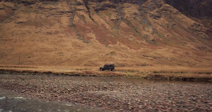 Flying over river tracking car driving through mountainous landscape. Panning up to reveal huge mountains