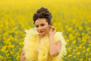 Portrait of romantic brunette teen girl wearing at yellow dress posing at field with yellow rapeseed flowers