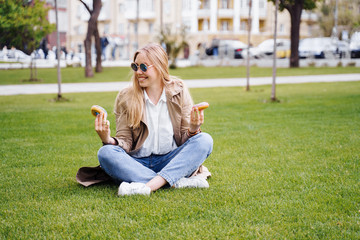 Beautiful, attractive, young caucasian girl with long blond hair sitting on the grass in a spring park. Woman holding donuts in her hands. Smiling and having a good time in nature outdoors. Posing