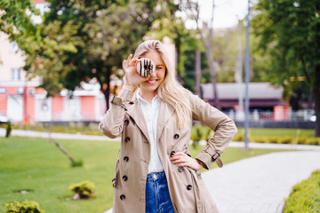 Beautiful, attractive blonde girl in a beige cloak and blue jeans is fooling around with donuts in her hands. Woman holding donuts near the face instead of eyes and smiling broadly.  Looking through