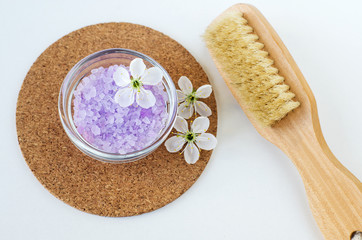 Small glass bowl with purple bath salt (foot soak), massage body brush and white flowers. Top view, copy space.
