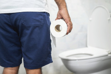 Man suffers from diarrhea hand hold tissue paper roll in front of toilet bowl. constipation in...