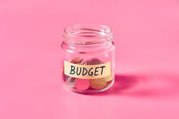 Glass jar with coins and inscription budget lies on pink background