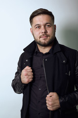 Young Bearded Dark Haired Man In Black Stylish Shirt, Jacket On Gray Background, Confident Male Holds Hands On Edge Clothing And Poses For Camera. Concept Of The Top Models