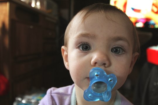 Close-up Portrait Of Cute Baby Girl Sulking Pacifier At Home