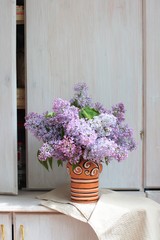 A bouquet of lilacs in a rustic ceramic vase on country wooden sideboard with an open door. Sun light