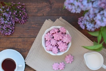 Small violet merengues in a plate. Cup of tea and milk in bottom corners.   bouquet of lilacs in blur, a lilac branch lays in a corner. Dark wooden table
