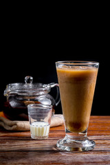 Iced milk tea in tall glass with solf focus of transparent teapot with hot tea on brown cloth on wooden table on black background with copy space. Famous beverage of Asian. Healthy drink concept