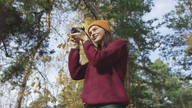 Beautiful young woman photographer dressed in knitted red jumper and yellow hat taking photos of sun in autumn forest. Low angle 4K 360 degree tracking arc shot.