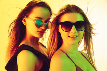 Beautiful young girls' portrait isolated on yellow studio background in neon light. Women in fashionable bodysuits. Facial expression, summer, weekend, beauty, resort concept. Vacations, youth.