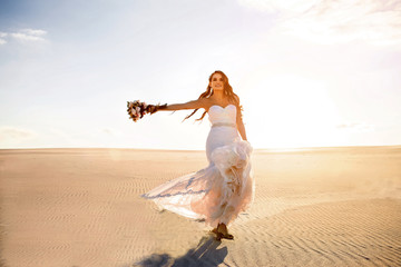 Fototapeta na wymiar a beautiful smiling bride in a white wedding dress walks along the sandy desert, a brunette with a wedding bouquet spins and has fun, sunset, sunshine