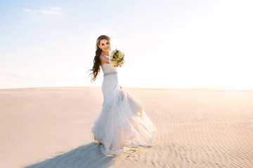 beautiful smiling bride in a white wedding dress walks in the sandy desert, a brunette girl with a wedding bouquet is spinning and having fun, sunset
