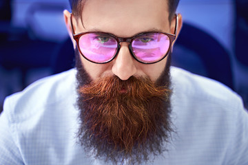 Bearded man in glasses with screen reflection working on computer in IT office, sitting at desk...