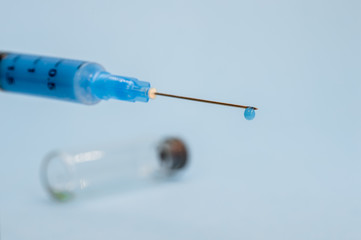 A close-up syringe needle with a drop of solution at the end against an empty vial on a blue background