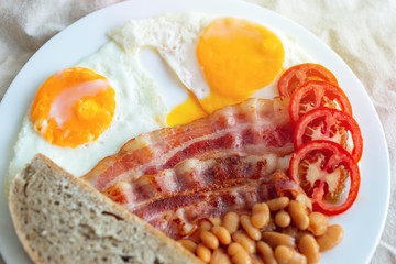 Close up of traditional English breakfast: fried eggs, bacon, beans, toast bread and tomato on a plate.