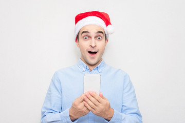 Amazed guy in Santa Claus hat with phone, portrait, white background. Concept of a new year.