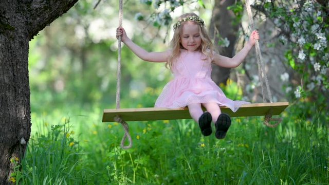 SLOW MOTION Happy cute little girl swinging on a swing against the background of a blossoming apple tree. Girl in a pink dress in a blooming garden. Happy childhood concept.