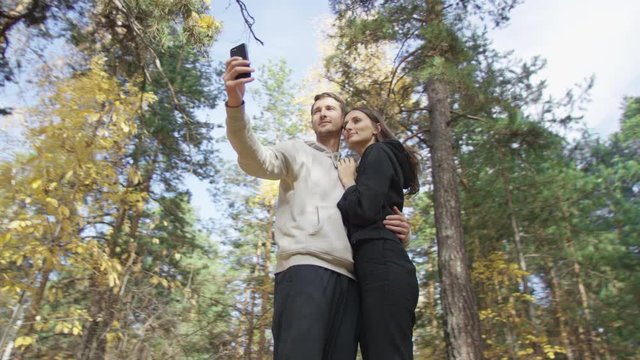 Enamoured couple dressed in sportive clothes taking selfie together with autumn forest at the background. Low angle 4K 360 degree tracking arc shot.