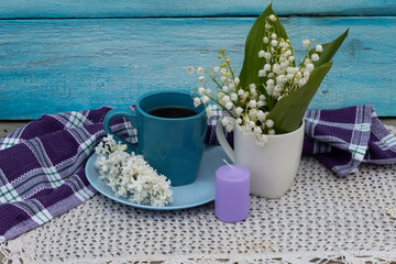 On a blue background on a white knitted napkin is a cup of tea, on a saucer is a white lilac. Nearby there is a cup with lilies of the valley and a candle.