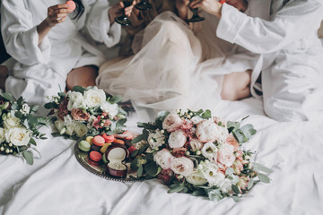 Modern wedding bouquets, wedding rings and delicious macarons on white bed on background of bride and bridesmaids celebrating. Wedding preparations or bridal shower.