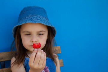 A smiling girl in a headdress 4-5 years old eats fresh strawberries in the open air. Summer season. Childhood. Isolated blue background