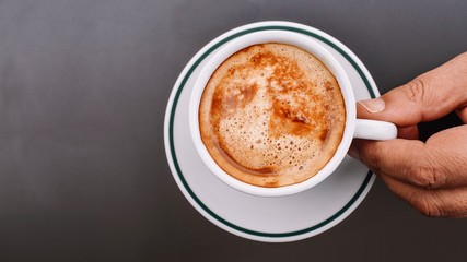 Close-up top view of a male hand holding a cup of creamy white coffee, ready to drink, on a gray background with copy space