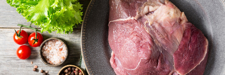Beef leg in a ceramic plate on the gray wooden kitchen table. Raw beef meat. A large piece of meat lies on a plate next to a wooden bowl with pepper, lettuce, and a knife. Banner with space for text