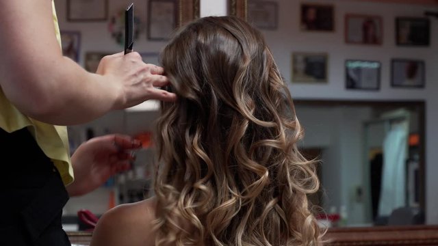 A master collects many curls to an evening hairstyle for a model with long dark-blond hair. Close-up shot, view of the nape.