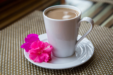 
White cup with aromatic coffee with milk on a white saucer with a pink flower for decoration.