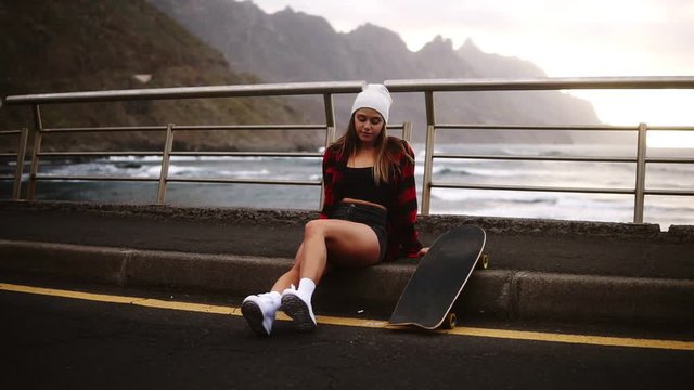 Girl sitting with longboard at the border at coastline asphalt road on background hills and foggy ocean. Relaxing break. Cloudy weather