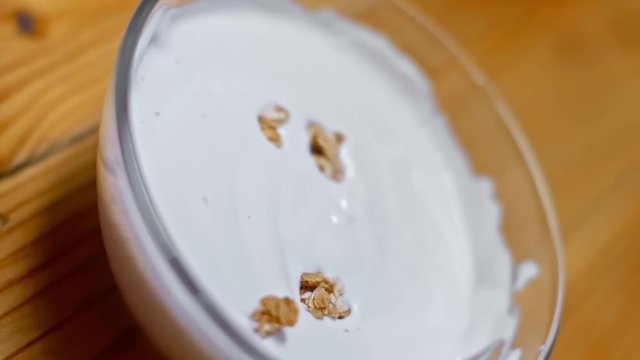 Granola fall into yogurt in a glass plate on a wooden table. Healthy breakfast or snacks, clean food concept. Slow motion