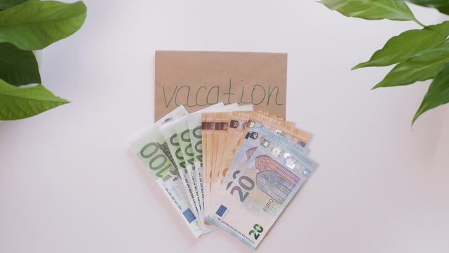 Top down view. Female hands count european money, euro, and then put it on an envelope with an inscription, the word Vacation. Background is a white table with green plants in the corners.