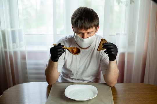 COVID-19. Young Caucasian Man In A White Mask And Black Gloves With Slice Of Bread, Wooden Spoon And A Bowl Of Soup Of Water At A Table In The Kitchen During A Pandemic. Coronavirus In Europe.