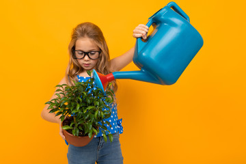 blond girl watering a leaf plant from a watering can on a yellow background