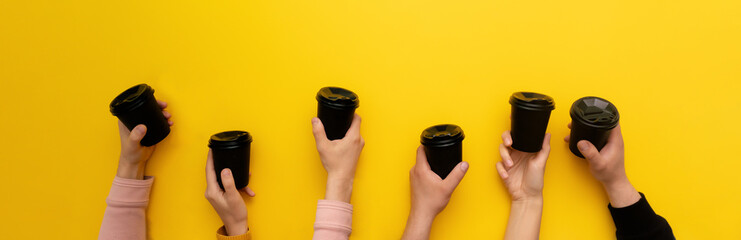 Black takeaway coffee cups with black lids in many caucasian woman and man hands on yellow background, banner, copy space.