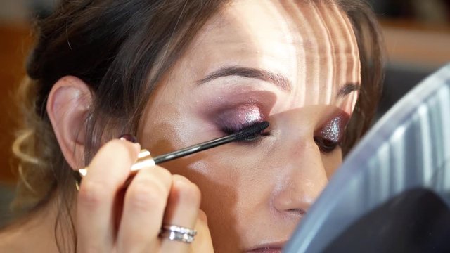 A beautiful model with evening hairstyle and make-up draws herself eyelashes with a black mascara. The model is looking in the mirror. 