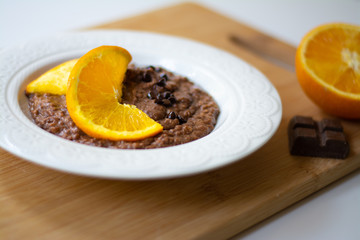 A bowl of healthy breakfast chocolate orange oatmeal porridge with chocolate chips and coconut flakes sprinkled on it. Orange and chocolate in the background. Stylish breakfast food concept.