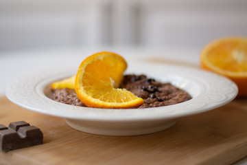A bowl of healthy breakfast chocolate orange oatmeal porridge with chocolate chips and coconut flakes sprinkled on it. Orange and chocolate in the background. Stylish breakfast food concept.