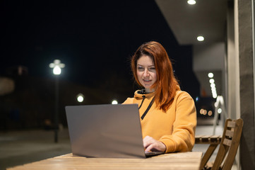 A woman works remotely at a laptop in a summer cafe late in the evening. Happy Girl studying while sitting on an empty street at a wooden table. Female freelancer in a sweatshirt at the evening
