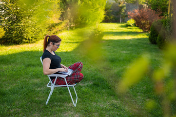 A woman with glasses sits in a folding chair with her legs crossed in a lotus position and typing on a laptop. Female freelancer remotely works in the garden of the cottage during quarantine.