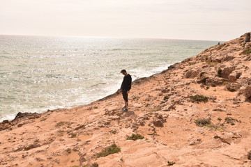 Young boy looking to the sea on a cliff with a black t-shirt. Looks like desert, or maybe not. 