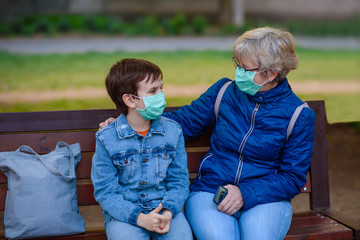 Fototapeta na wymiar Boy 10 years old outdoors is sitting on a park bench with grandmother and talking. Pensioner woman with kid in protective masks during quarantine. Covid-19, pandemic coronavirus 2020.