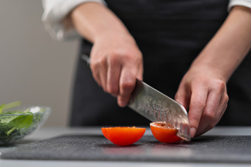 A female chef in a white uniform and a black apron in the restaurant kitchen. The cook cuts the vegetables with a large kitchen knife.