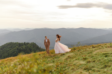 The bride and groom are standing close to each other against the backdrop of autumn mountains. The groom admires his wife.